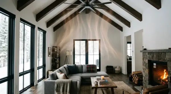Salon cosy avec cheminée en pierre, canapé gris, grandes fenêtres donnant sur la neige. Le **chauffage au plafond** discret assure une chaleur douce.