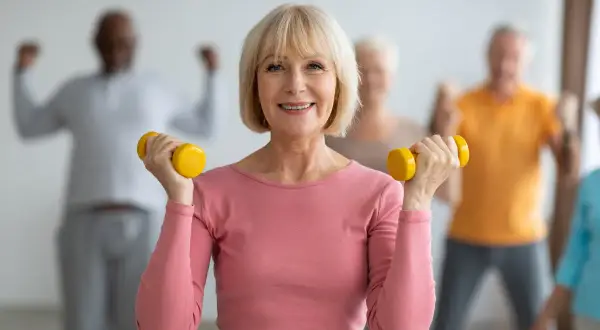 Une femme souriante fait de la gymnastique senior avec des haltères jaunes. D'autres personnes âgées s'entraînent en arrière-plan.