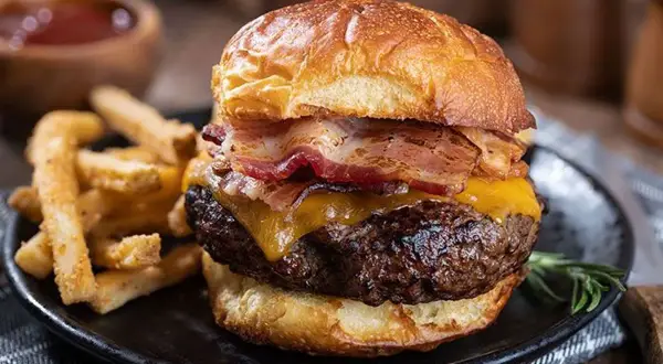 Gros plan sur un burger maison avec bacon, fromage fondant et frites croustillantes sur une assiette noire.