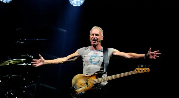 Sting śpiewa na koncercie, trzymając gitarę basową. Jego ramiona są rozłożone, jakby chciał objąć cały świat. Tekst "sting shape of my heart" idealnie pasuje do tej sceny.