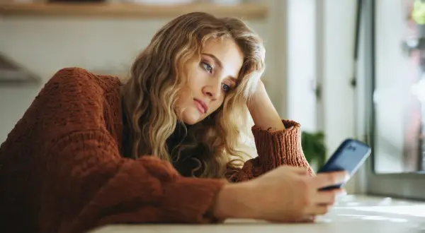 Młoda kobieta w brązowym swetrze, z ręką opartą o głowę, patrzy na telefon. Jej wyraz twarzy sugeruje anhedonię, brak radości z życia.