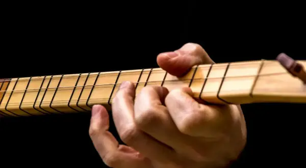 Dłoń gra na instrumencie, dociskając struny. Widoczne są progi i nazwy strun w gitarze.