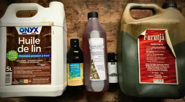 Plusieurs bouteilles d'huile pour le bois, dont de l'huile de lin et du goudron de pin, sont alignées sur une surface en bois.