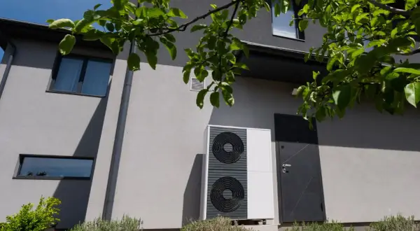 Unità esterna di una pompa di calore residenziale, moderna e discreta, installata accanto a una porta d'ingresso su un edificio grigio.
