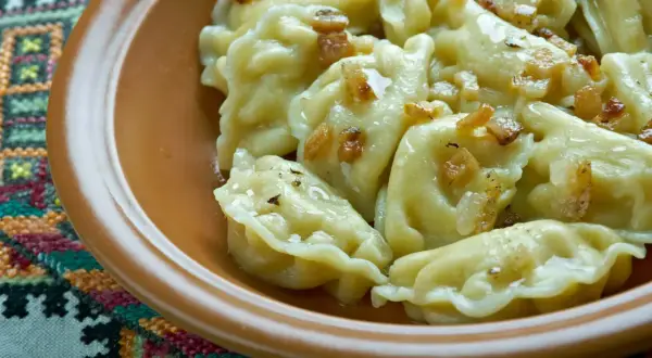 Pyszne pierogi ruskie z podsmażoną cebulką i skwarkami, podane w glinianej misce na haftowanej serwetce.