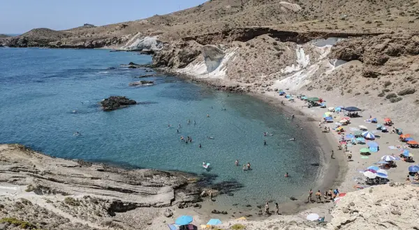Bahía de aguas cristalinas con gente bañándose y disfrutando de las **cabo de gata playas**. Sombrillas de colores y arena dorada bajo un cielo azul.