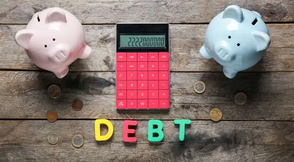 Due salvadanai rosa e azzurro, una calcolatrice rossa con un numero elevato e lettere che formano la parola "DEBT" suggeriscono un **debito residuo** da gestire.