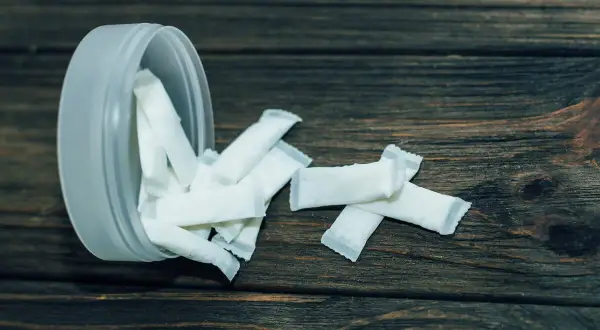 Białe woreczki z nikotyną, popularne jako snus zęby, rozsypane obok plastikowego pojemnika na drewnianym tle.