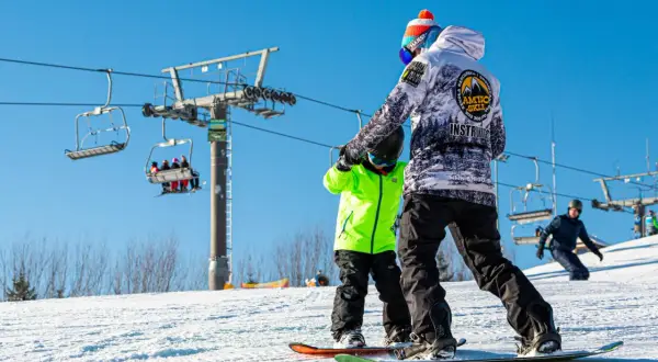 Instruktor snowboardingu uczy dziecko na stoku, w tle kolejka krzesełkowa.