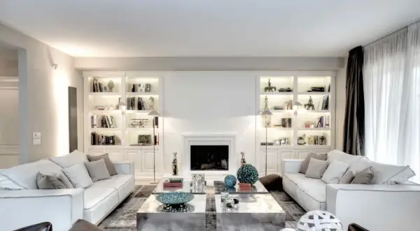 Soggiorno elegante con divani bianchi, librerie illuminate e camino. Le pareti color bianco latte creano un'atmosfera luminosa e ariosa.