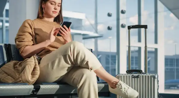 Junge Frau mit Handy in der Airport Lounge, Koffer daneben.
