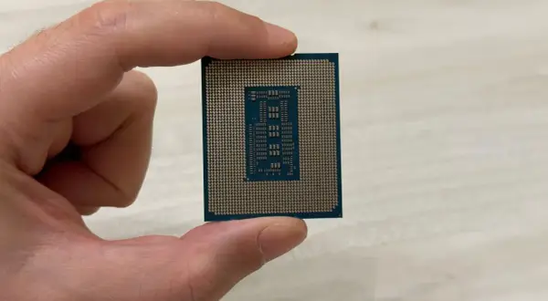 Un dedo sostiene un procesador o CPU, mostrando su parte inferior con pines dorados y un diseño rectangular en el centro.