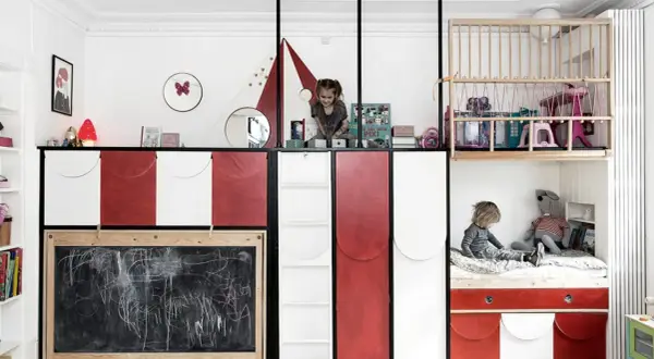 Zwei Kinder teilen sich ein Zimmer mit kreativen Schlafkojen und Spielbereichen. Ein Kind sitzt auf einem Bett, das andere spielt oben.