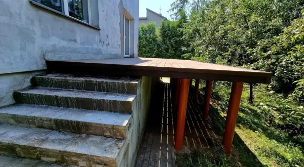 Nowy taras na słupach stalowych, z drewnianą podłogą, sąsiaduje z betonowymi schodami i zielonym ogrodem.