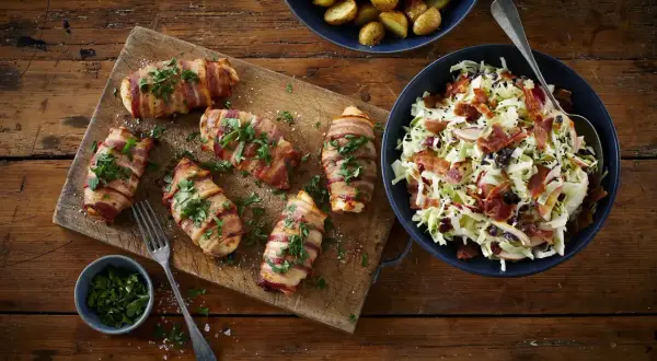 Sex saftiga baconlindad kycklingar på en skärbräda, serverade med en fräsch coleslaw och rostade potatisar.
