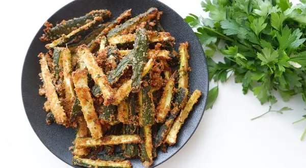 Zucchine gratinate in padella, dorate e croccanti, servite su un piatto nero con prezzemolo fresco.