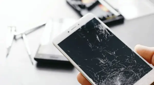 Dłoń trzyma telefon z pękniętym ekranem. Obok leżą narzędzia do naprawy.