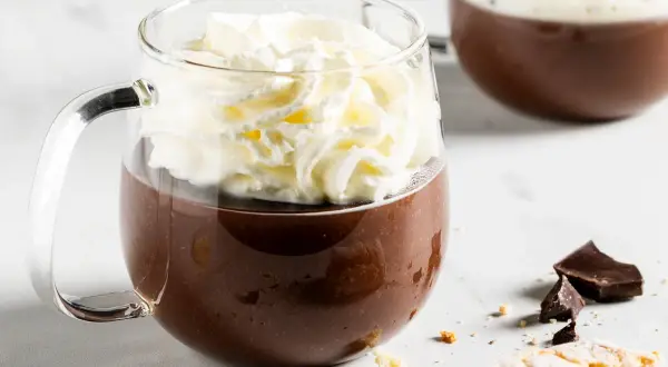 Una tazza di fumante cioccolata calda coronata da panna montata, con pezzi di cioccolato e biscotti sullo sfondo.