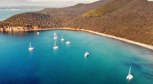 Vela al largo della costa della Maremma Toscana. Il mare azzurro e cristallino abbraccia una baia con diverse barche a vela e un catamarano.