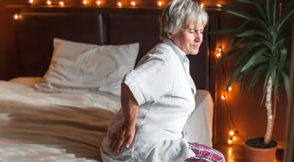 Elderly woman grimaces in pain, holding her hip. She looks like she slept on her hip wrong, sitting on a bed with festive lights.