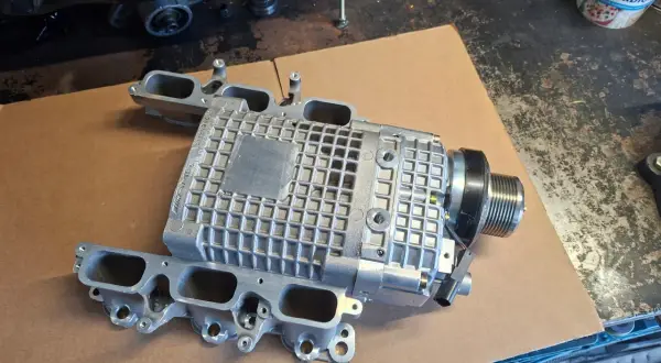 Srebrna sprężarka mechaniczna z kolektorem dolotowym, część do Mercedes Benz C32 AMG, leży na kartonie.