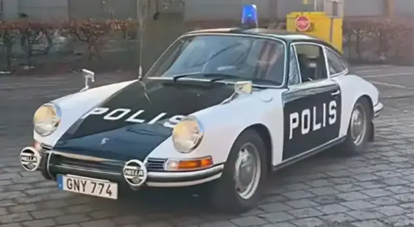 En klassisk Porsche polisbil med blåljus på taket. Bilen är vit med svarta detaljer och texten "POLIS" på motorhuven och dörren.
