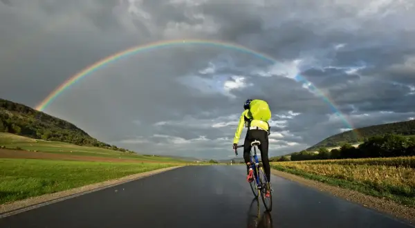 Rennradtour 3 Tage: Ein Radfahrer fährt unter einem Regenbogen auf nasser Straße.