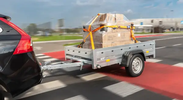 Nowy projekt przyczepki samochodowej UNITRAILER w akcji. Pudełka zabezpieczone pasami na przyczepce ciągniętej przez czarne auto.