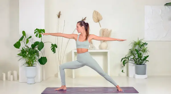 Mujer en postura de Virabhadrasana 2, con brazos extendidos y mirada al frente, rodeada de plantas y decoración minimalista.