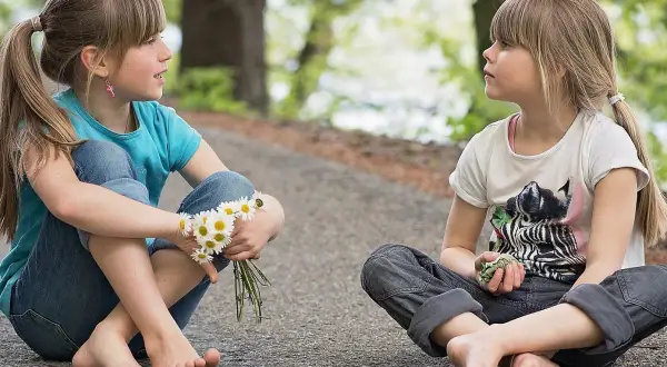 Dwie dziewczynki siedzą na ścieżce, rozmawiając. Jedna trzyma stokrotki. To inspiracja, jak nauczyć dziecko asertywności.