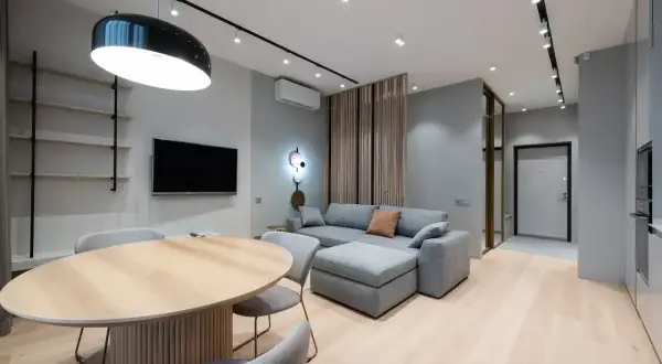 Nowoczesny salon z szarą kanapą i drewnianym stołem. Ściany w odcieniu cool white, jasna podłoga.