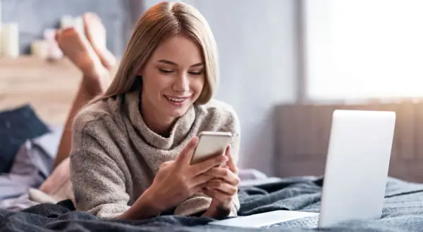 Uśmiechnięta dziewczyna leży na łóżku z telefonem w dłoniach, szukając informacji, jak doładować komuś konto w Play z karty na kartę. Obok leży laptop.