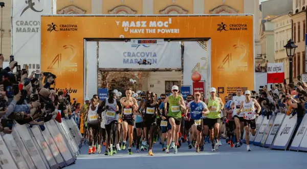Biegacze startują w maratonie. Wielka brama z napisem "Cracovia Maraton" i hasłem "Masz moc!" symbolizuje wyzwanie, ile ma maraton.