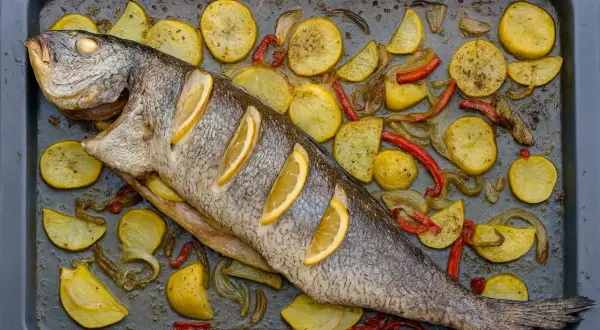 Corvina al horno con patatas, pimientos y rodajas de limón en una bandeja.