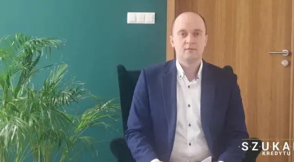 Mężczyzna w garniturze siedzi w fotelu obok rośliny, na tle zielonej ściany i drzwi.