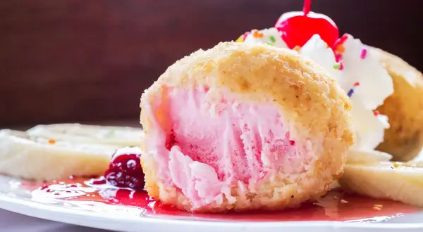 Ein knusprig gebratenes Eis mit rosa Füllung, garniert mit Sahne, Kirsche und bunten Streuseln. Dazu Bananenscheiben und rote Sauce.