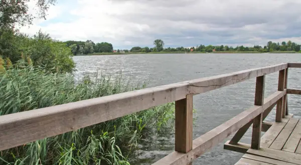 Drewniany pomost nad jeziorem, gdzie można łowić ryby. W oddali widać zalesiony brzeg i domy.