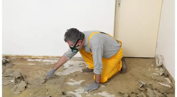 Un homme en salopette jaune et masque retire la colle de moquette avec une spatule. Une méthode naturelle pour enlever colle moquette.