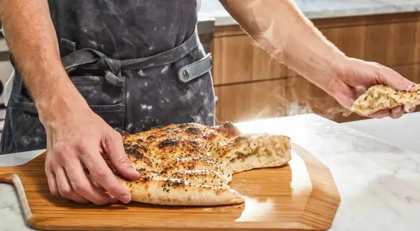 En person i förkläde håller upp en bit av en nybakad, ångande ramazan pidesi. Brödet är gyllene med sesamfrön.
