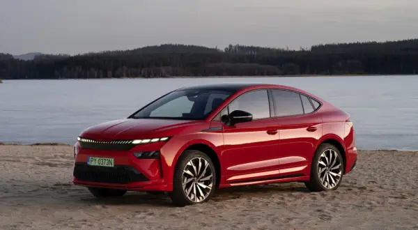 Czerwony Skoda Enyaq na piaszczystym brzegu, z lodowym jeziorem i lasem w tle.
