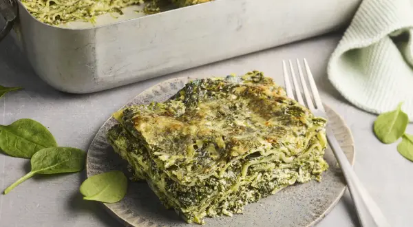 Eine Portion Spinat Ricotta Lasagne auf einem Teller, daneben eine Auflaufform mit der restlichen Lasagne. Frische Spinatblätter liegen daneben.