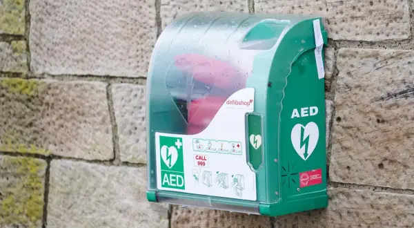 Boîtier vert fixé sur un mur de pierre, contenant un défibrillateur. Des pictogrammes indiquent qui peut utiliser un défibrillateur et comment l'utiliser.
