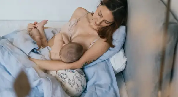 Matka w wygodnej pozycji biologicznej do karmienia, przytulona do poduszki, karmi piersią swoje dziecko.