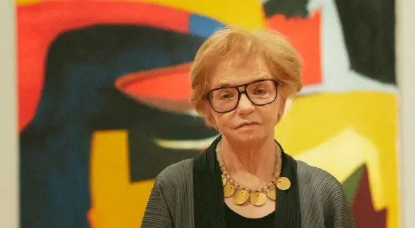 A woman with glasses and a statement necklace stands before a vibrant abstract expressionist painting.