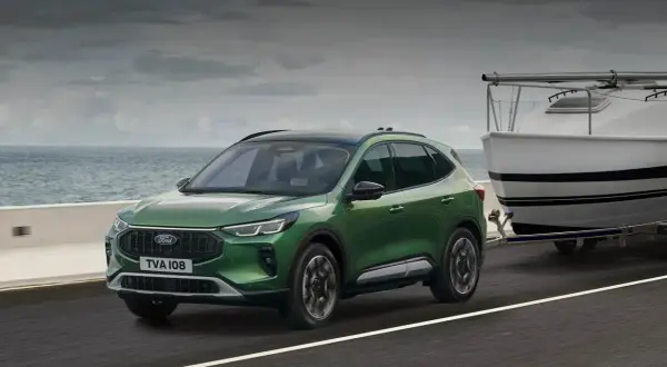 Zielony Ford Kuga z przyczepą i łodzią nad morzem. Sprawdź aktualną ford kuga cena.