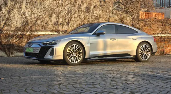 Srebrny Audi e-tron GT lśni w słońcu na brukowanej ulicy, z pnączami na ścianie w tle.