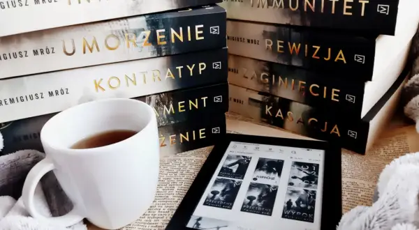 Stos książek Remigiusza Mroza, biały kubek z herbatą i czytnik Kindle.