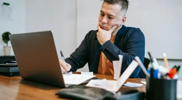 Zamyślony audytor siedzi przy biurku, pisząc coś na papierze i patrząc na ekran laptopa.