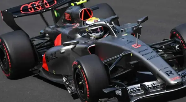 Un coche de **Audi F1** gris y negro con detalles rojos acelera en la pista. El piloto lleva casco amarillo y blanco.