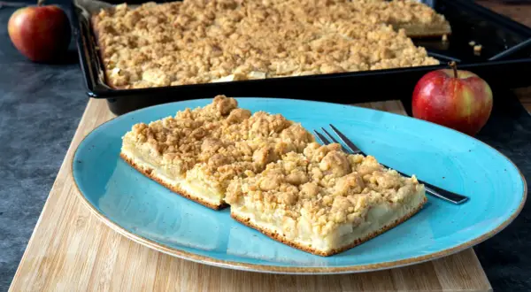 Zwei Stücke saftiger Apfelkuchen mit Streuseln auf einem blauen Teller, daneben ein ganzer Apfel. Perfekt für ein Apfelkuchen Rezept.